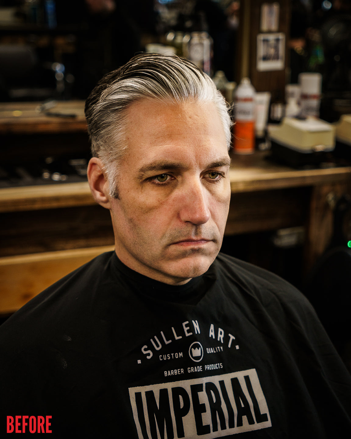 Before shot of a man wearing a barber cape in a barbershop, silver hair loosely slicked back prior to styling with Imperial Blacktop Pomade.