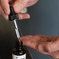 A tattooed hand draws the dropper from a 1 oz bottle of Imperial Pre-Shave Oil and Beard Conditioner held over an open palm.