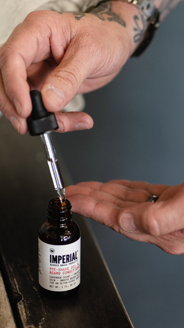 A tattooed hand draws the dropper from a 1 oz bottle of Imperial Pre-Shave Oil and Beard Conditioner held over an open palm.