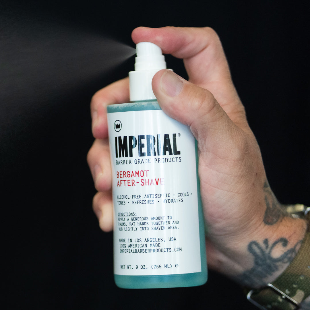 A tattooed hand pressing the spray pump of a 9 oz Imperial Bergamot After-Shave bottle, with mist visible against a dark background.