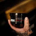 A tattooed hand holding a 6 oz black tub of Imperial Blacktop Pomade in a barbershop setting.