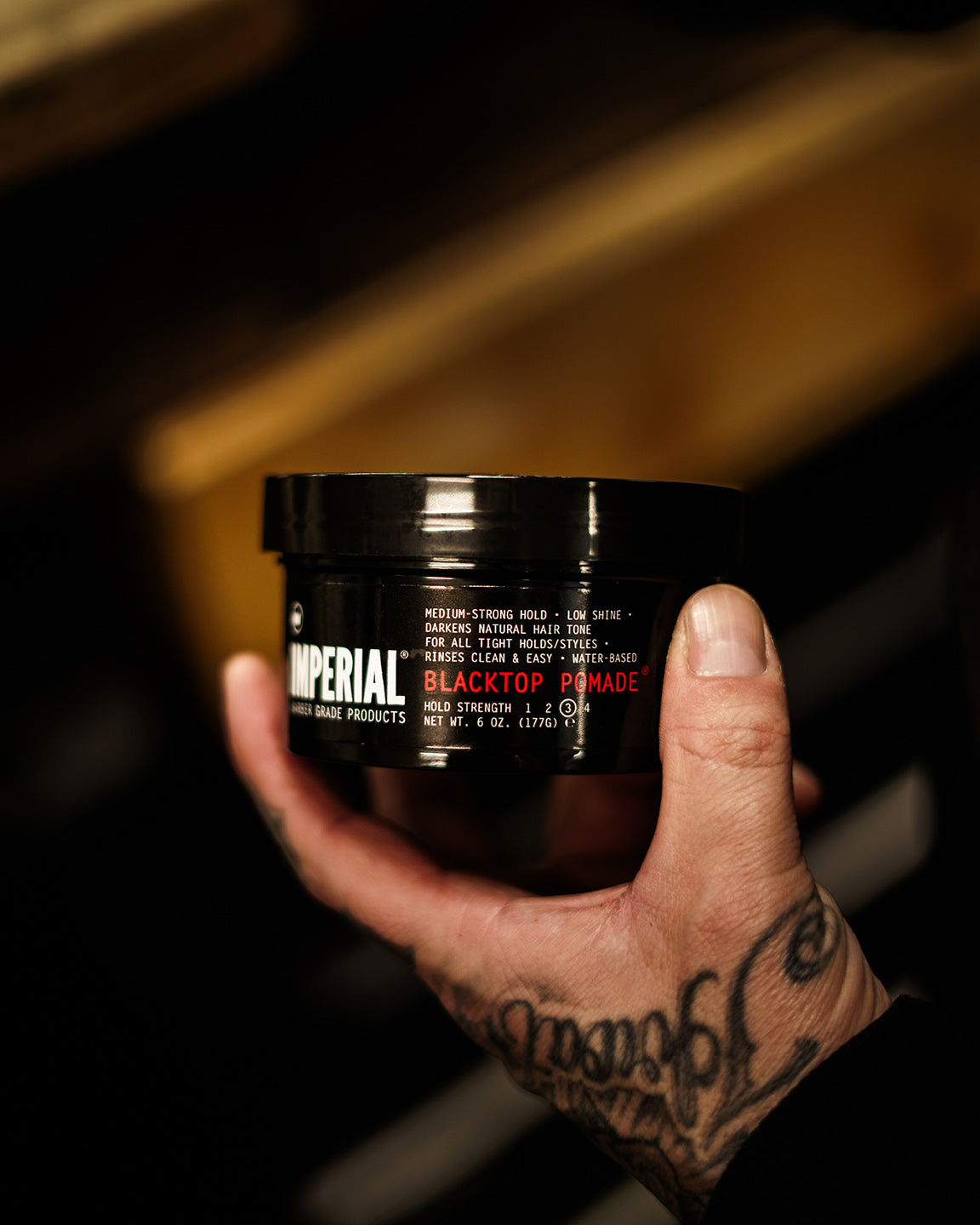 A tattooed hand holding a 6 oz black tub of Imperial Blacktop Pomade in a barbershop setting.