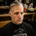 Before shot of a man wearing a barber cape in a barbershop, silver hair loosely slicked back prior to styling with Imperial Blacktop Pomade.