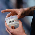 A tattooed hand opens the clear 6 oz tub of Imperial Classic Pomade, revealing the product inside.