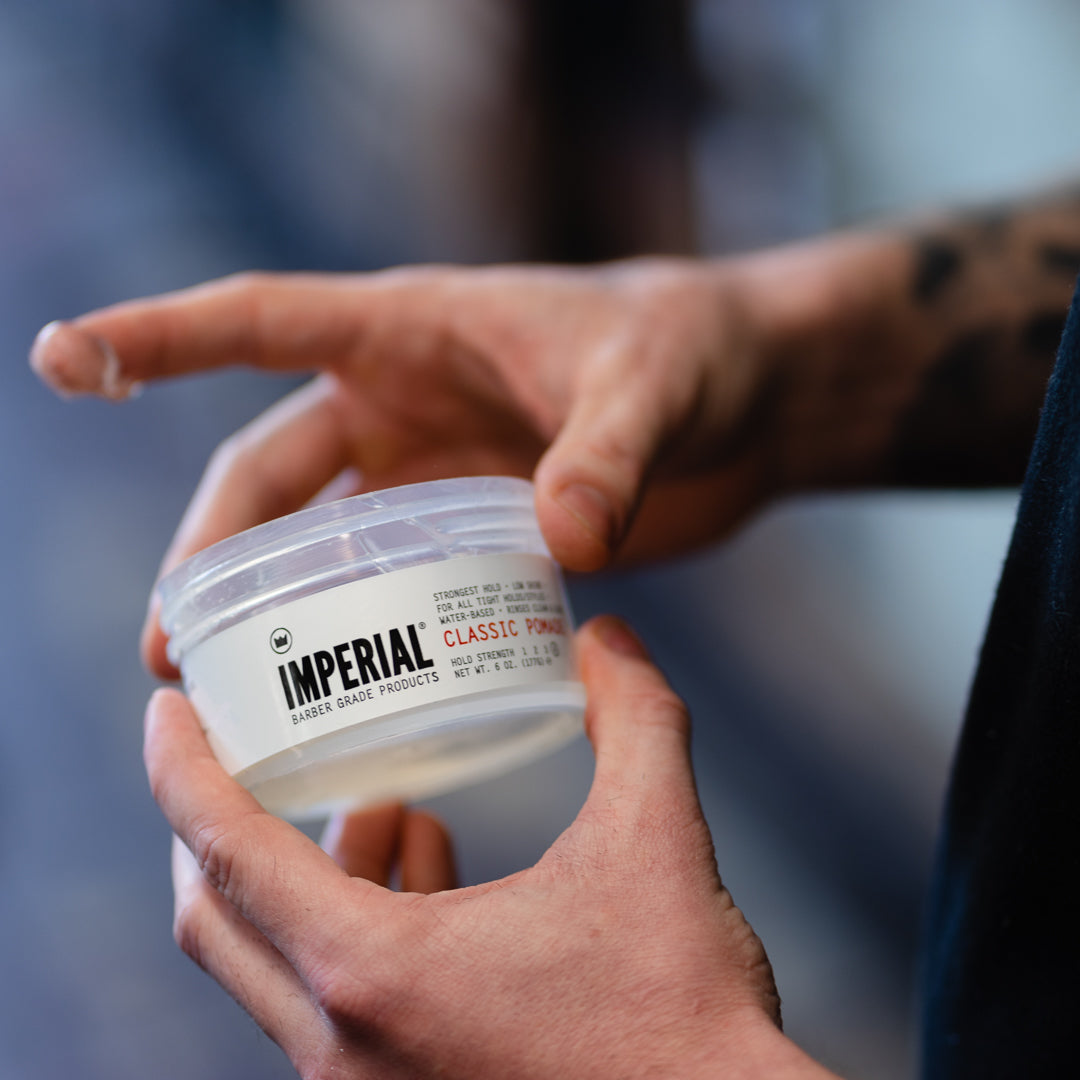 A tattooed hand opens the clear 6 oz tub of Imperial Classic Pomade, revealing the product inside.