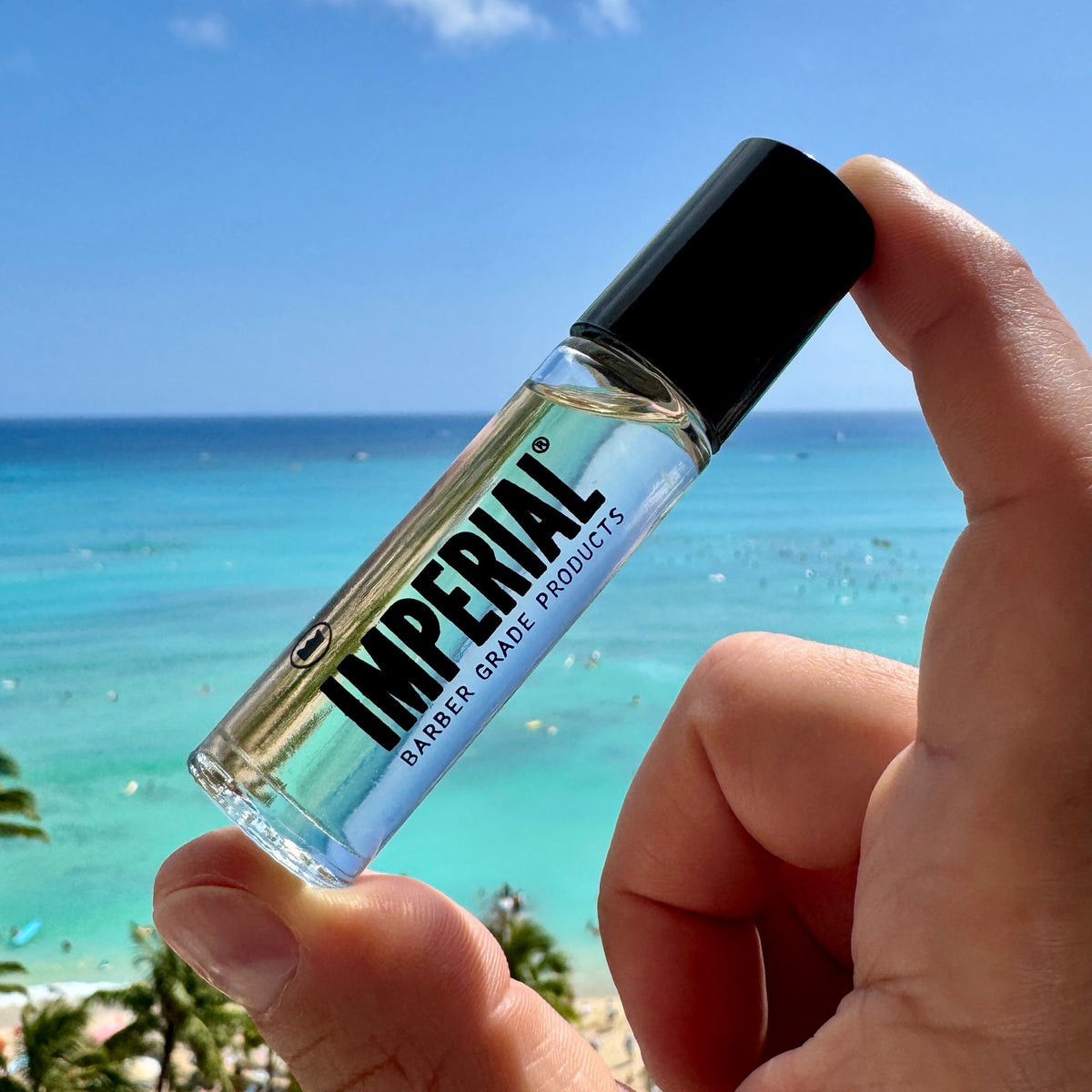 A hand holds the Imperial Barber Grade Products Crown Roll cologne bottle against a bright turquoise ocean backdrop.