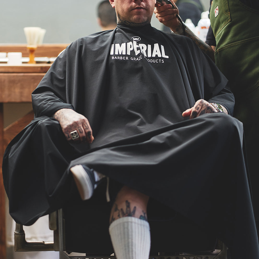 A tattooed man seated in a barber chair wearing the black Imperial Barber Grade Products cape while a barber works on his fade.