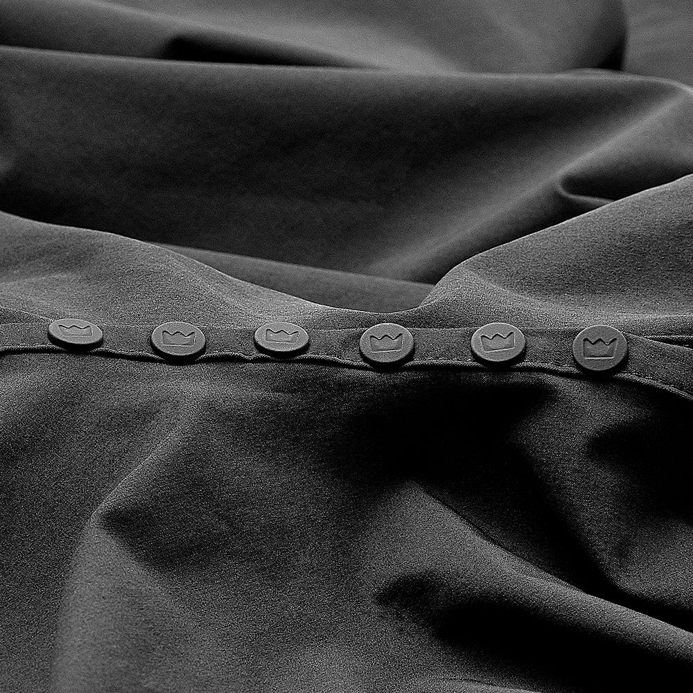 Close-up of the Imperial Barber Cape Standard Edition neckline, showing a row of seven dark snap buttons each embossed with the Imperial crown logo.