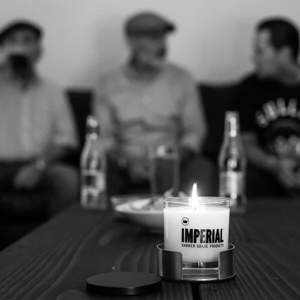Imperial Barber Grade Products Standard Candle burning on a wood table in the foreground, with three men socialising in the blurred background.