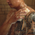 A tattooed man scrubs his shoulder with a luffa sponge in the shower, soap lather covering his back and neck.