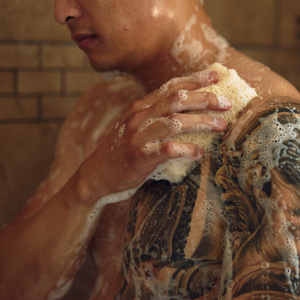 A tattooed man scrubs his shoulder with a luffa sponge in the shower, soap lather covering his back and neck.